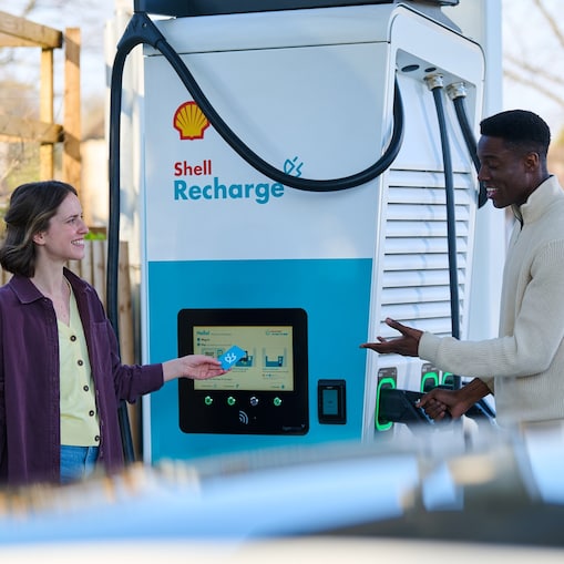 Eine Person hält eine Shell Recharge Ladekarte vor das Kartenlesegerät einer E-Ladestation, während eine weitere Person den Ladestecker aus der Ladestation nimmt.