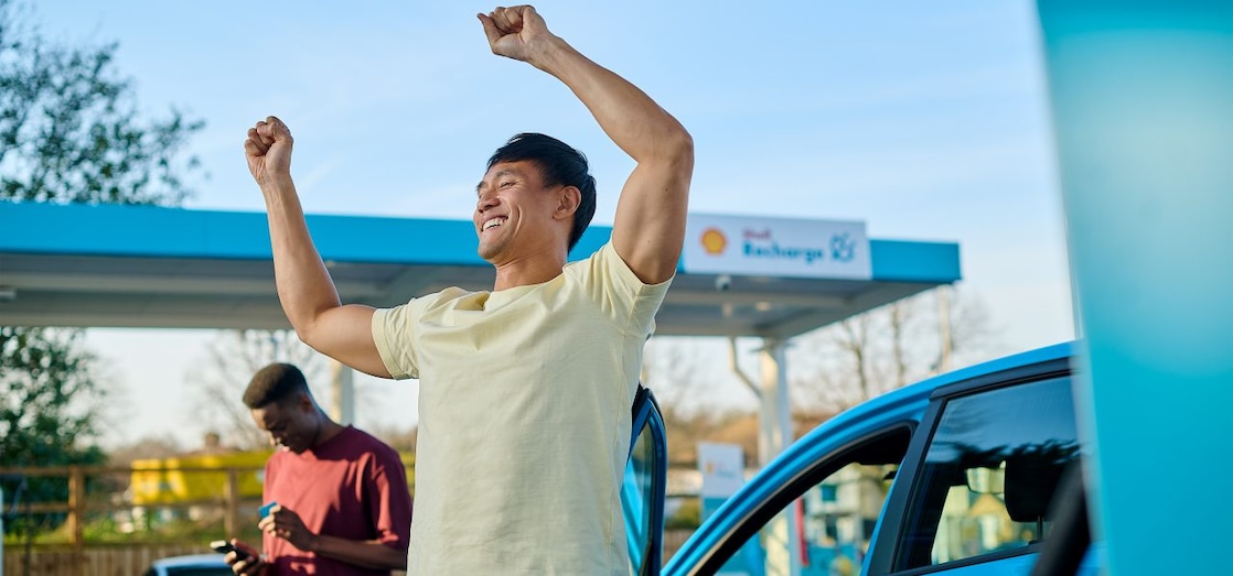Eine Person steht neben einem Elektroauto mit geöffneter Fahrzeugtür und hebt die Arme. Im Hintergrund befindet sich eine Shell Recharge E-Ladestation, während eine weitere Person neben dem Fahrzeug auf ein Smartphone blickt.