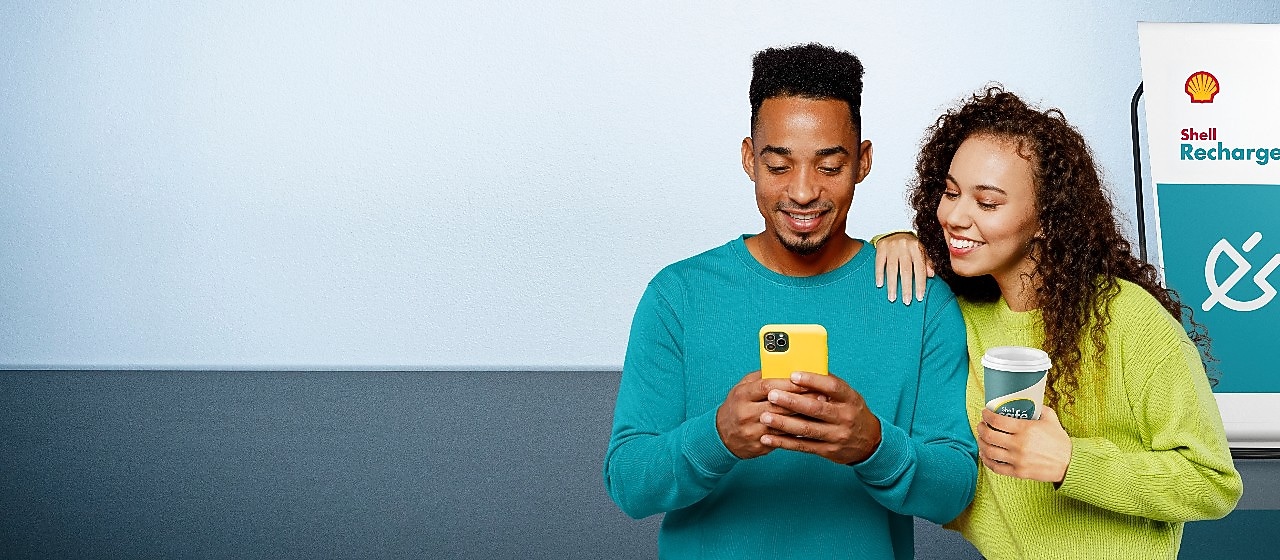 Ein lächelndes Paar steht nebeneinander; der Mann hält ein gelbes Smartphone, die Frau lehnt sich an ihn und hält einen Kaffeebecher. Im Hintergrund ist eine Shell Recharge Ladesäule zu sehen.