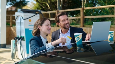 Eine Frau und ein Mann nutzen das Autodach als Arbeitsfläche und schauen gemeinsam auf einen Laptop. Daneben stehen zwei Shell Café Becher; im Hintergrund ist eine Shell Recharge Ladesäule zu sehen.