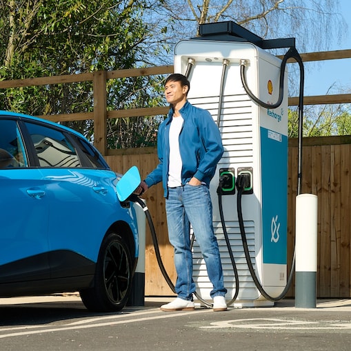 Eine Person steht neben einem Elektrofahrzeug an einer Shell Recharge Schnellladesäule. Das Fahrzeug ist mit der Ladesäule verbunden und wird geladen.