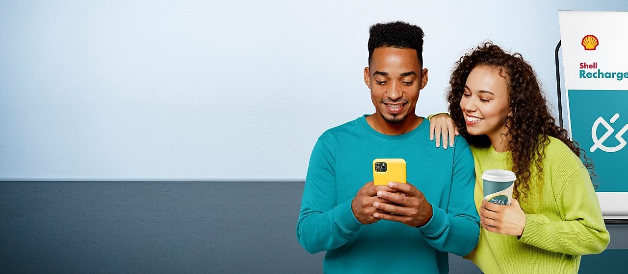 Ein lächelndes Paar steht nebeneinander; der Mann hält ein gelbes Smartphone, die Frau lehnt sich an ihn und hält einen Kaffeebecher. Im Hintergrund ist eine Shell Recharge Ladesäule zu sehen.