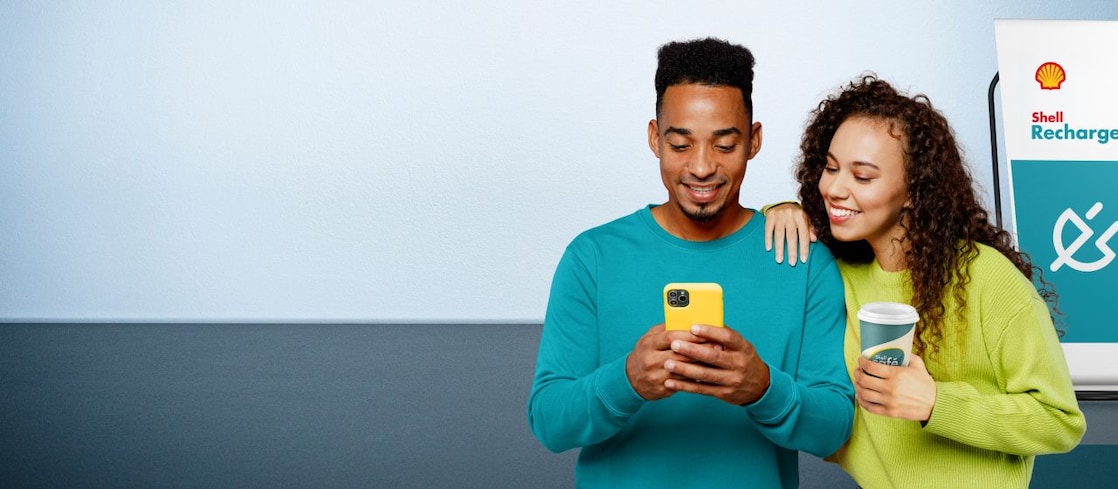 Ein lächelndes Paar steht nebeneinander; der Mann hält ein gelbes Smartphone, die Frau lehnt sich an ihn und hält einen Kaffeebecher. Im Hintergrund ist eine Shell Recharge Ladesäule zu sehen.