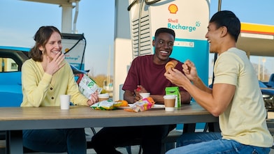 Drei Personen verbringen eine Pause an einer Shell Recharge E-Ladestation. Sie sitzen an einem Tisch mit Getränken und Snacks, während ein blaues Elektrofahrzeug im Hintergrund geladen wird.