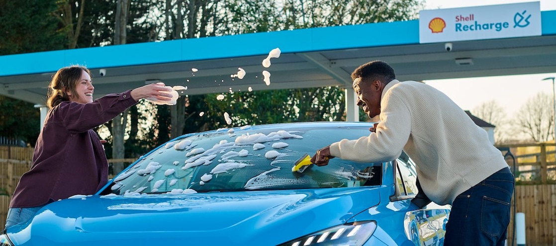 Zwei Personen reinigen ein blaues Elektrofahrzeug an einer Shell Recharge E-Ladestation. Eine Person wischt die Windschutzscheibe mit einem Schwamm ab, während die andere Person Seifenschaum über das Fahrzeug verteilt. Im Hintergrund sind die Shell Recharge Ladestation und weitere Ladepunkte sichtbar.