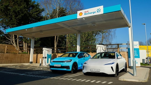 Zwei Elektroautos (blau und weiß) laden unter einem überdachten Shell Recharge-Schnellladepunkt. Im Hintergrund ein Holzzaun, Bäume und klarer Himmel an einem sonnigen Tag.