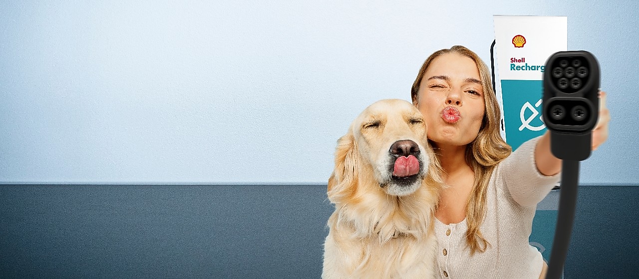 Eine Frau und ein Hund strecken spielerisch die Zunge bzw. machen einen Kussmund, während sie ein Ladekabel vor einer Shell-Recharge-Ladestation in die Kamera halten.