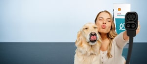 Eine Frau und ein Hund strecken spielerisch die Zunge bzw. machen einen Kussmund, während sie ein Ladekabel vor einer Shell-Recharge-Ladestation in die Kamera halten.