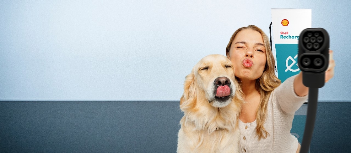 Eine Frau und ein Hund strecken spielerisch die Zunge bzw. machen einen Kussmund, während sie ein Ladekabel vor einer Shell-Recharge-Ladestation in die Kamera halten.