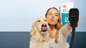 Eine Frau und ein Hund strecken spielerisch die Zunge bzw. machen einen Kussmund, während sie ein Ladekabel vor einer Shell-Recharge-Ladestation in die Kamera halten.