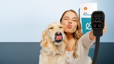 Eine Frau und ein Hund strecken spielerisch die Zunge bzw. machen einen Kussmund, während sie ein Ladekabel vor einer Shell-Recharge-Ladestation in die Kamera halten.