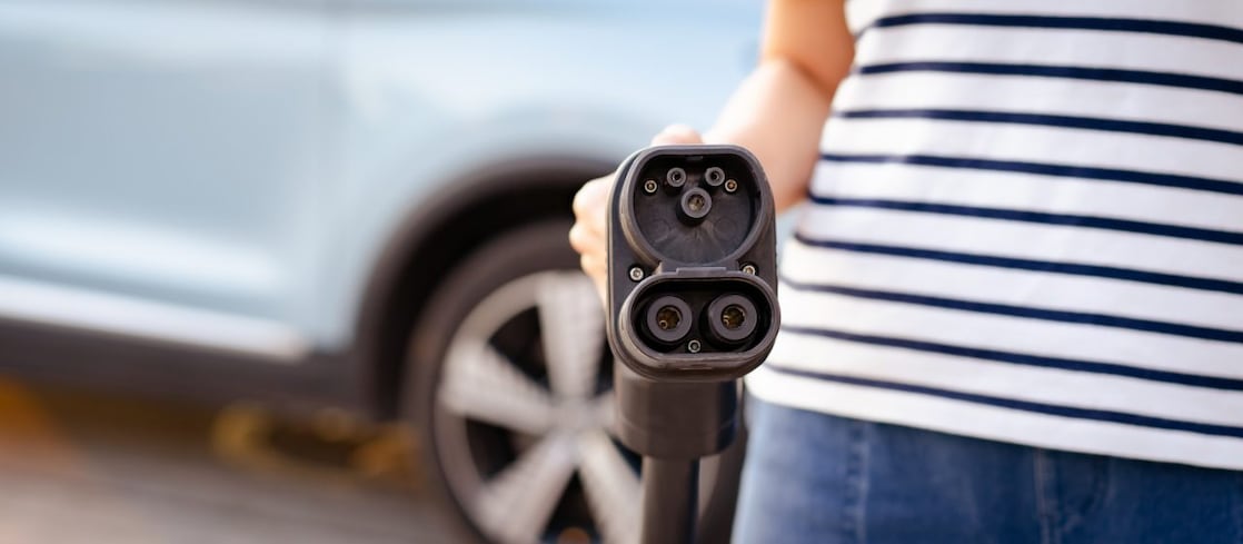  Eine Person hält ein Ladekabel für ein Elektroauto direkt zur Kamera, im Hintergrund steht ein hellblaues Auto.