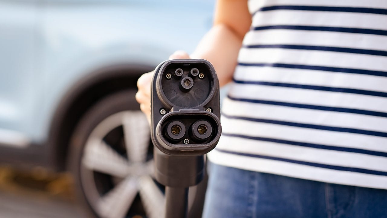  Eine Person hält ein Ladekabel für ein Elektroauto direkt zur Kamera, im Hintergrund steht ein hellblaues Auto.