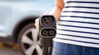  Eine Person hält ein Ladekabel für ein Elektroauto direkt zur Kamera, im Hintergrund steht ein hellblaues Auto.