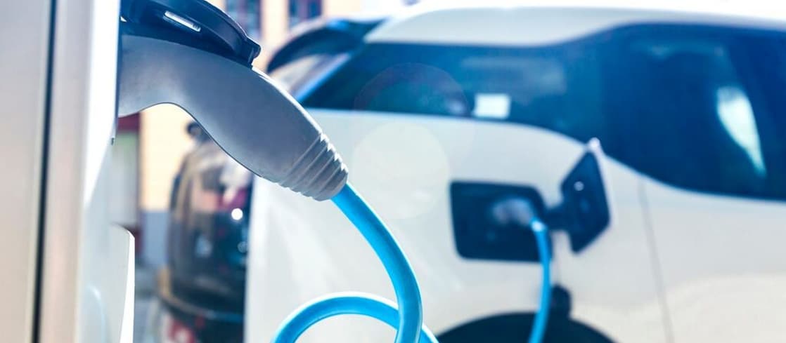 Nahaufnahme eines blauen Ladekabels, das in ein weißes Elektroauto eingesteckt ist; im Hintergrund sind weitere Fahrzeuge zu sehen.