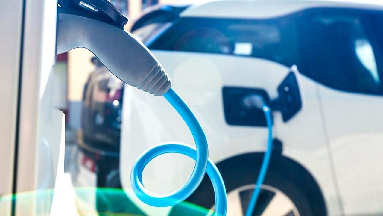  Nahaufnahme eines blauen Ladekabels, das in ein weißes Elektroauto eingesteckt ist; im Hintergrund sind weitere Fahrzeuge zu sehen.