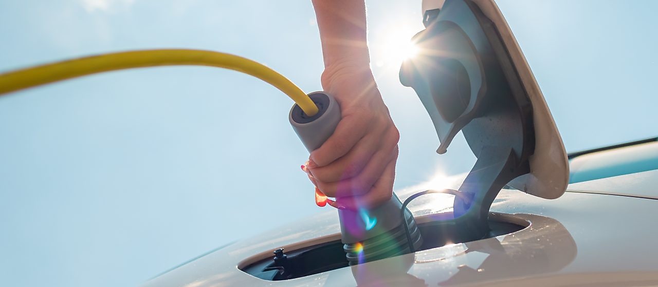 Eine Hand steckt bei starkem Sonnenlicht ein gelbes Ladekabel in den Ladeanschluss eines Elektroautos.