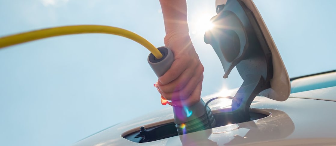  Eine Hand steckt bei starkem Sonnenlicht ein gelbes Ladekabel in den Ladeanschluss eines Elektroautos.