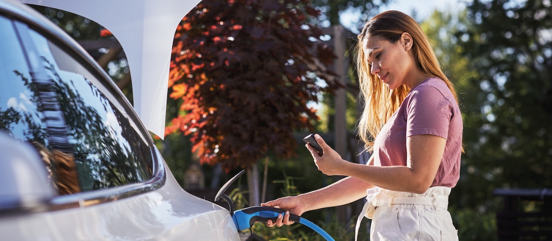  Eine Frau steckt im Freien ein blaues Ladekabel in ihr weißes Elektroauto und schaut dabei auf ihr Smartphone.