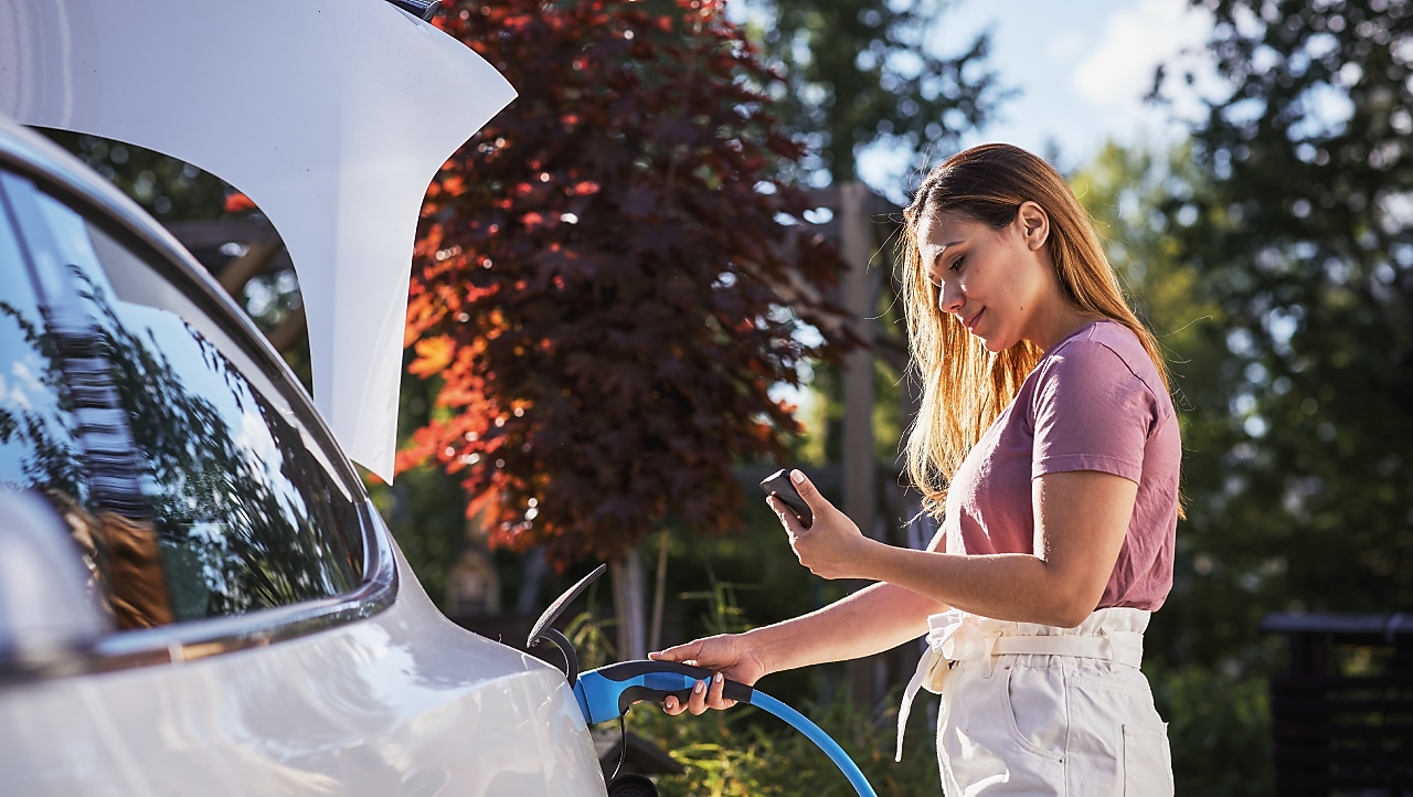  Eine Frau steckt im Freien ein blaues Ladekabel in ihr weißes Elektroauto und schaut dabei auf ihr Smartphone.