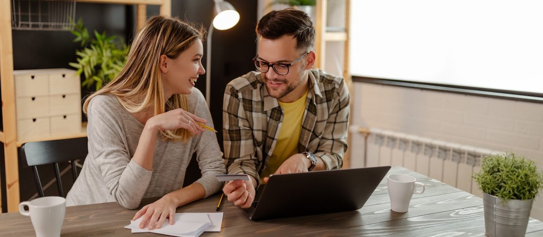  Ein Paar sitzt an einem Tisch, bespricht Unterlagen und schaut gemeinsam auf ein Smartphone; vor ihnen steht ein Laptop sowie Kaffee­tassen und eine Pflanze.