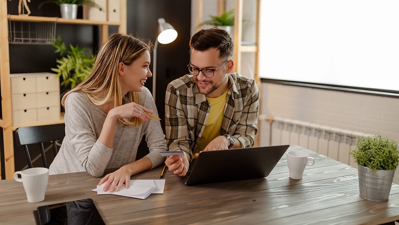  Ein Paar sitzt an einem Tisch, bespricht Unterlagen und schaut gemeinsam auf ein Smartphone; vor ihnen steht ein Laptop sowie Kaffee­tassen und eine Pflanze.