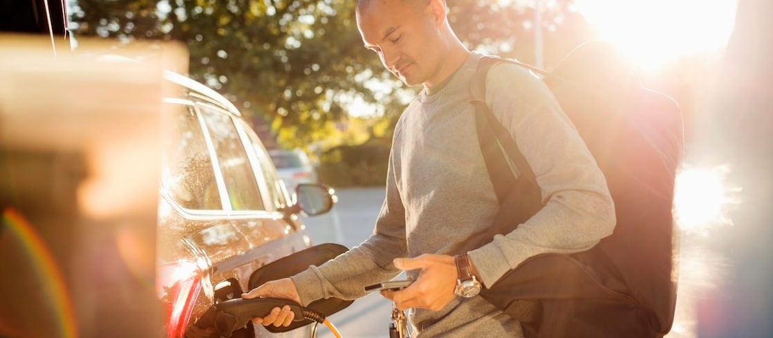 Ein Mann steckt bei tief stehender Sonne ein Ladekabel in sein geparktes Elektroauto und hält dabei sein Smartphone in der anderen Hand.