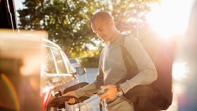 Ein Mann steckt bei tief stehender Sonne ein Ladekabel in sein geparktes Elektroauto und hält dabei sein Smartphone in der anderen Hand.