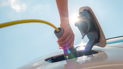 A female hand is holding a plug and charging an electric car, with a bright sun light and blue sky in the background