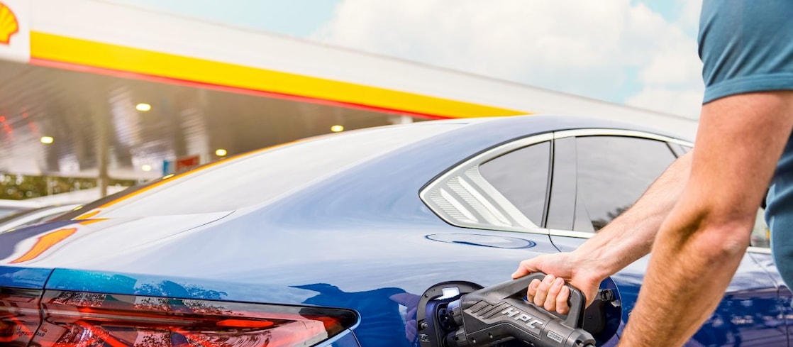  Ein Mann steckt ein Schnellladekabel in den Ladeanschluss eines blauen Elektroautos an einer Shell-Tankstelle.