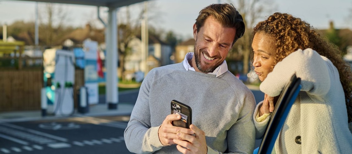 Ein Mann und eine Frau stehen lächelnd vor einer Shell Recharge Ladestation und schauen gemeinsam auf ein Smartphone. Im Hintergrund sind mehrere türkisfarbene Ladesäulen zu sehen.