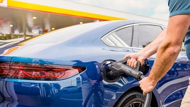 Ein Mann steckt ein Schnellladekabel in den Ladeanschluss eines blauen Elektroautos an einer Shell-Tankstelle.