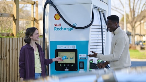 Eine Frau und ein Mann stehen vor einer Shell Recharge Ladesäule. Die Frau bedient das Display der Ladesäule. Im Hintergrund ist ein Fahrzeug zu sehen.