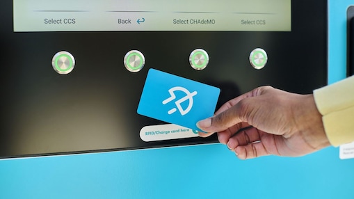 Eine Hand hält eine türkisfarbene Shell Recharge Ladekarte vor ein Kartenlesegerät einer Ladesäule. Auf dem Display im Hintergrund sind verfügbare Ladepunkte mit den Optionen CCS und CHAdeMO zu sehen.