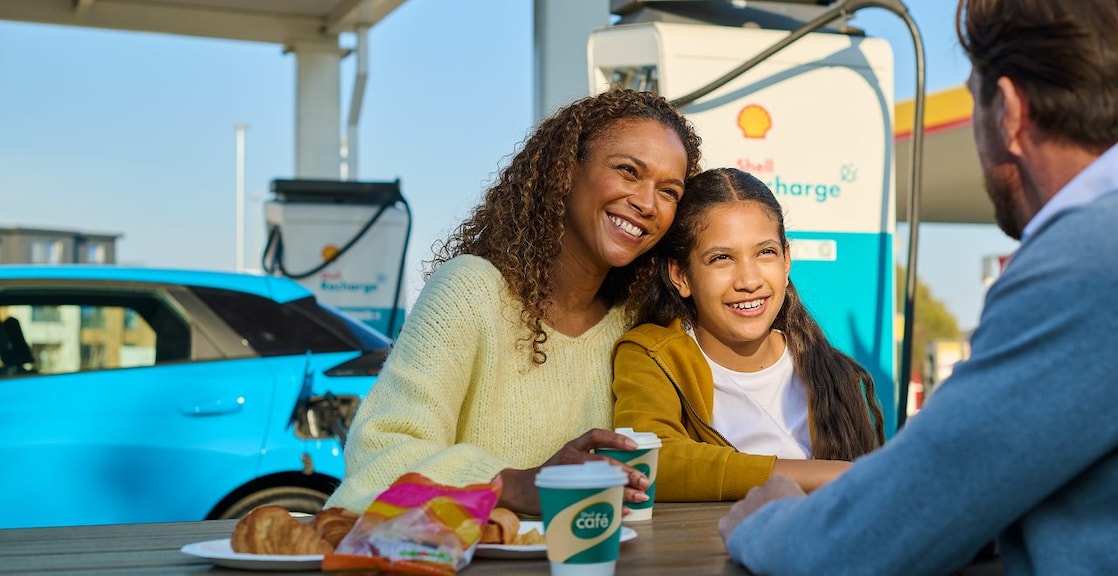 Personen machen eine Pause an einer Shell Recharge E-Ladestation. Im Hintergrund wird ein blaues Elektrofahrzeug geladen.