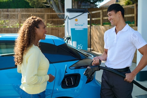 Zwei Personen stehen neben einem blauen Elektrofahrzeug an einer Shell Recharge E-Ladestation. Eine Person hält ein Ladekabel in der Hand.