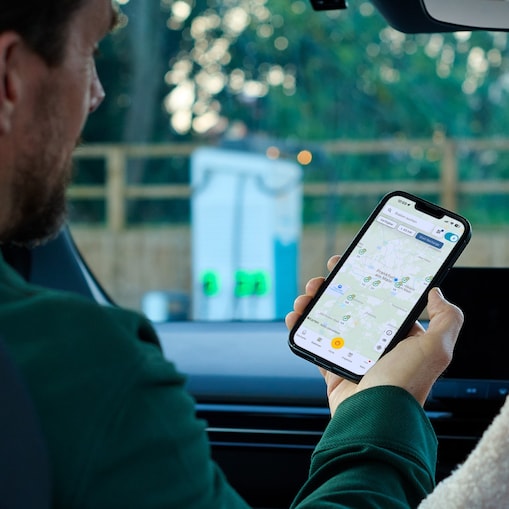 man-in-car-using-shell-app-station-finder Alt Text: Ein Mann sitzt im Auto und hält ein Smartphone mit der Shell-Recharge-App in der Hand. Die Karte zeigt verfügbare Ladestationen rund um Frankfurt am Main; draußen ist unscharf eine Ladesäule zu sehen.