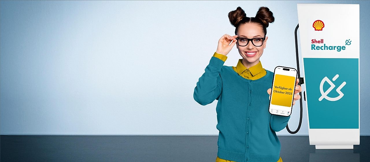 Eine lächelnde Frau mit Brille hält ein Smartphone hoch, auf dem „Verfügbar ab Oktober 2023“ steht; neben ihr steht eine Shell-Recharge-Ladestation.