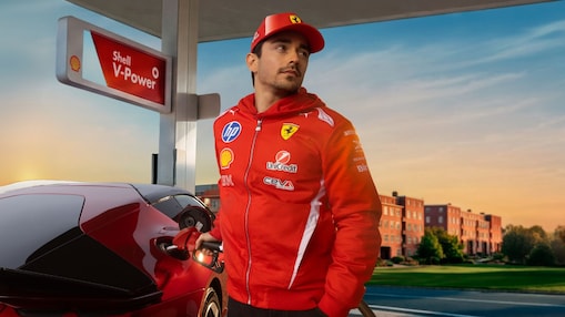 Formel-1-Fahrer Charles LeClerc in rotem Rennanzug betankt einen roten Sportwagen an einer Zapfsäule mit Shell V-Power Beschilderung bei warmem Abendlicht.