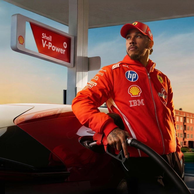 Formel-1-Fahrer Lewis Hamilton in rotem Rennanzug betankt einen roten Sportwagen an einer Zapfsäule mit Shell V-Power Beschilderung bei warmem Abendlicht.