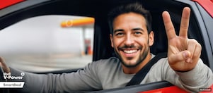 Ein Mann sitzt in einem roten Auto. Er ist angeschnallt und lächelt. Das Seitenfenster ist komplett unten, sodass er seinen Arm lässig hinaushält und mit seiner Hand das Victory-Zeichen macht. Im Hintergrund erkennen wir in der Unschärfe eine Shell Tankstelle.