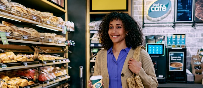  Frau steht lächelnd mit einem Kaffee von Shell Café in einer Shell Station mit Shell Café Backshop