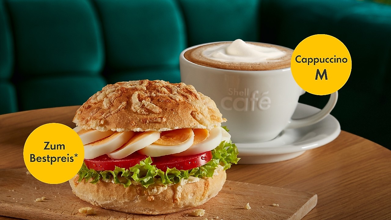 Auf einem Holztisch befinden sich ein belegtes Käsebrötchen mit Ei, Tomate und Salat auf einem Holzbrett mit einem gelben Störer mit dme Text "Zum Bestpreis*". Daneben ein Cappuccino mit gelben Störer mit Text "Größe M".