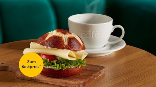 Ein Fußball-Laugenbrötchen belegt mit Gouda, Tomate, Gurke und Salat serviert auf einem kleinen Holzbrett. Im Hintergrund eine dampfende weiße Shell Café Tasse mit Untertasse auf einem Holztisch vor einem petrolfarben gepolsterten Sitz. Ein Störer in Form eines gelben Kreises im Vordergrund trägt die Aufschrift „Zum Bestpreis*“