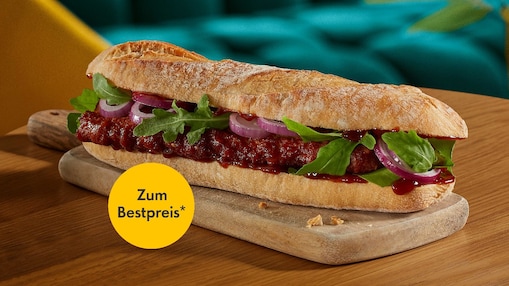 Ein Rustico-Baguette belegt mit einem Fleischpatty, BBQ-Sauce, Rucola und roten Zwiebeln serviert auf einem kleinen Holzbrett auf einem Holztisch. Im Hintergrund ein petrolfarben gepolsterter Sitz. Ein Störer in Form eines gelben Kreises im Vordergrund trägt die Aufschrift „Zum Bestpreis“